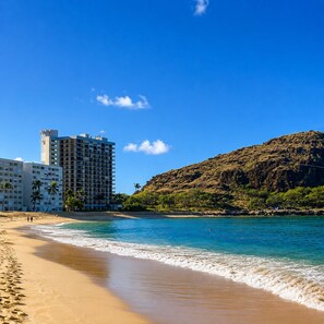 A beach with clear blue water, sandy shore, and a rocky hill in the background.