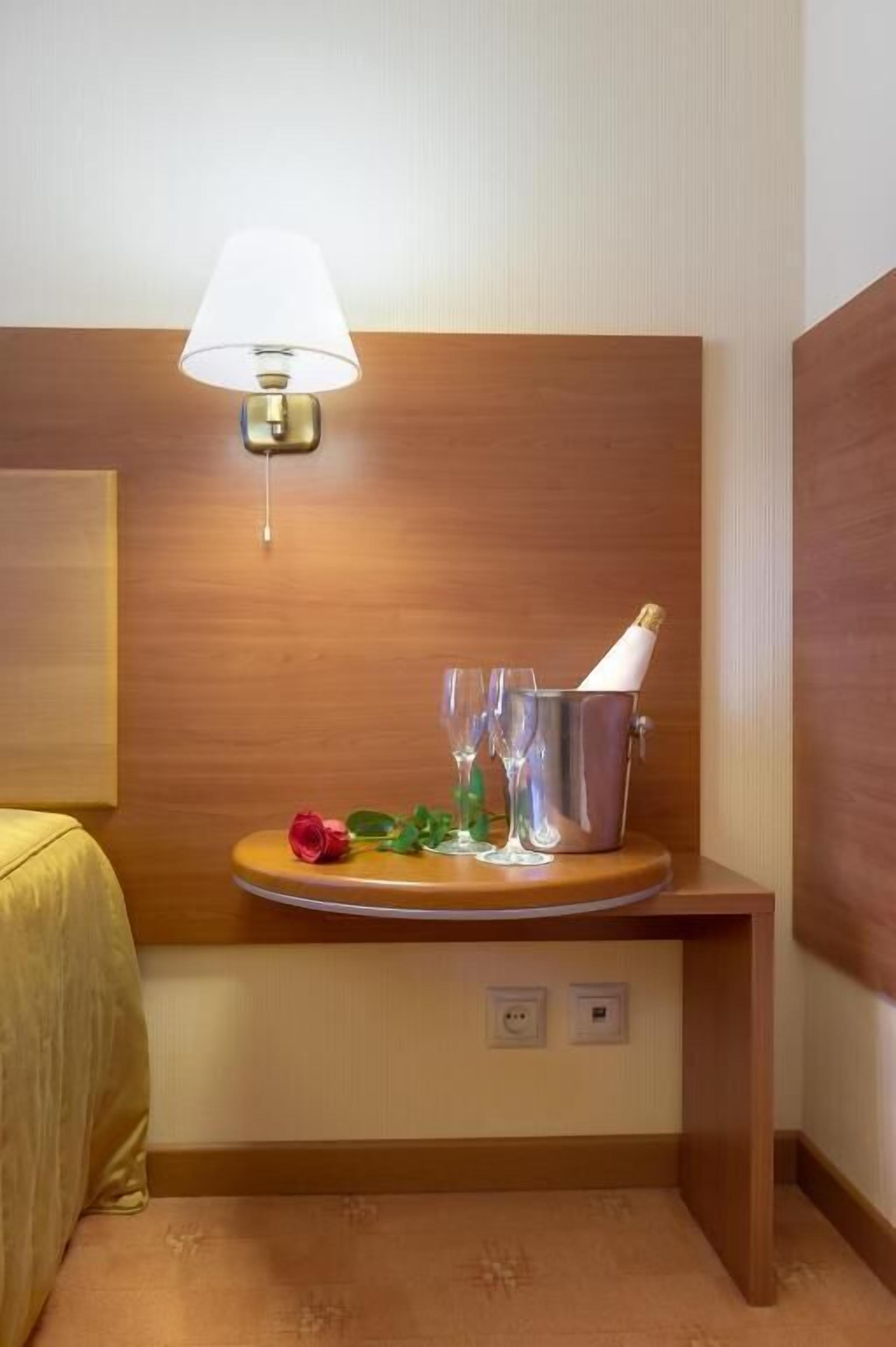A hotel room with a wooden headboard, a bedside table with a lamp, a small table with a champagne bottle, glasses, and a rose.
