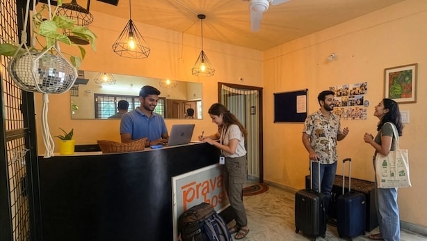 A reception area with a desk, a sign reading 'Prava Host,' and a wall with photos. There are plants hanging from the ceiling and a person at the desk.