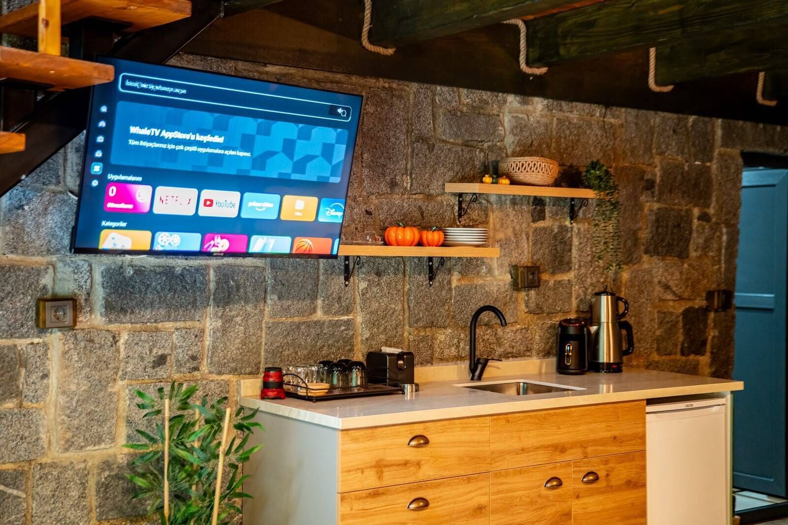 Una cocina con pared de piedra, estantes de madera, un lavamanos y un televisor montado mostrando una interfaz de YouTube.