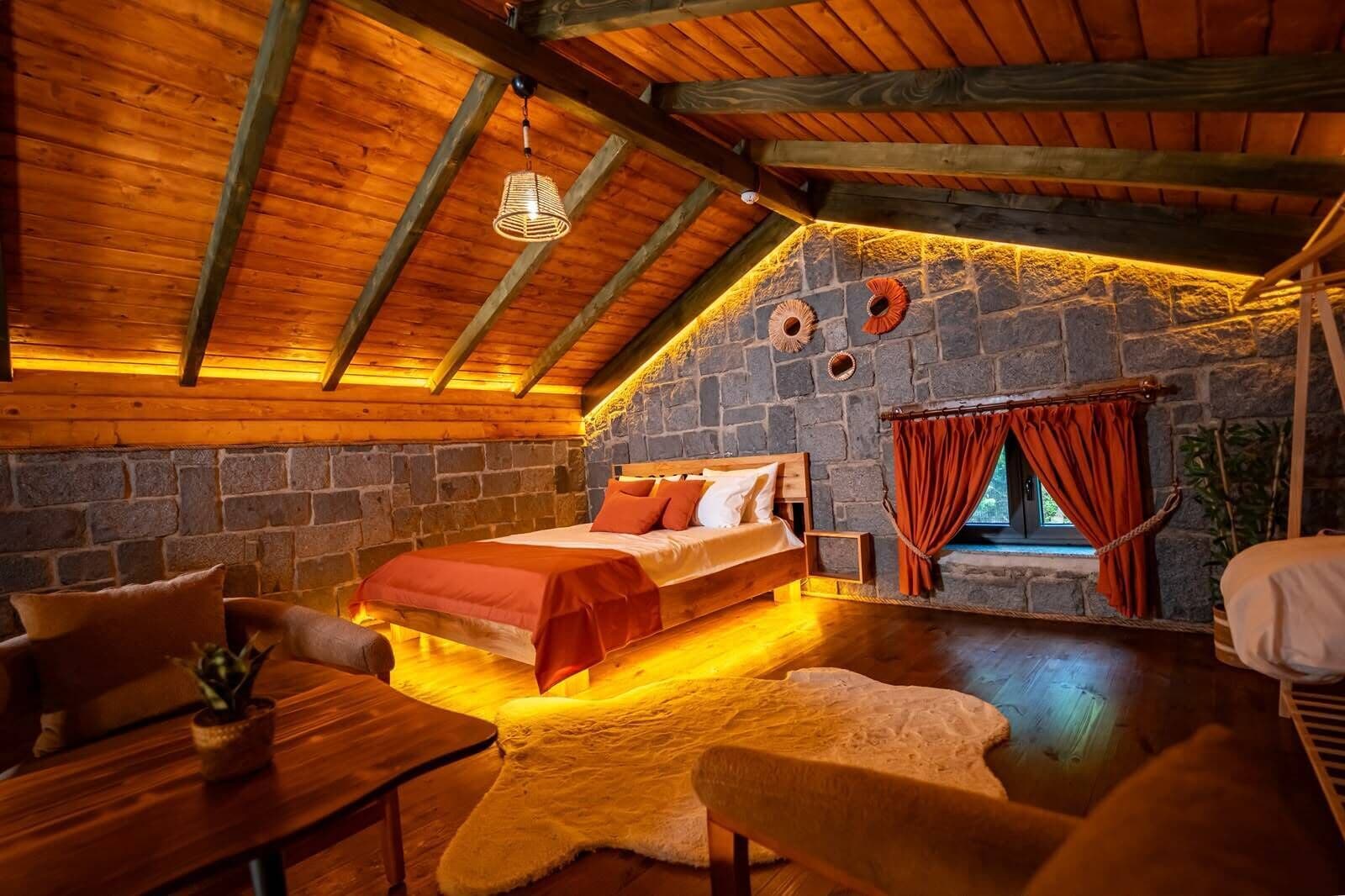 A cozy room with a bed, a wooden table, a chair, and a stone wall.