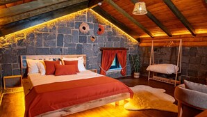A cozy attic bedroom with a stone wall, a wooden bed, a hanging chair, and a window with red curtains. - Dolana Wadi Taş Evleri (Ardeşen)