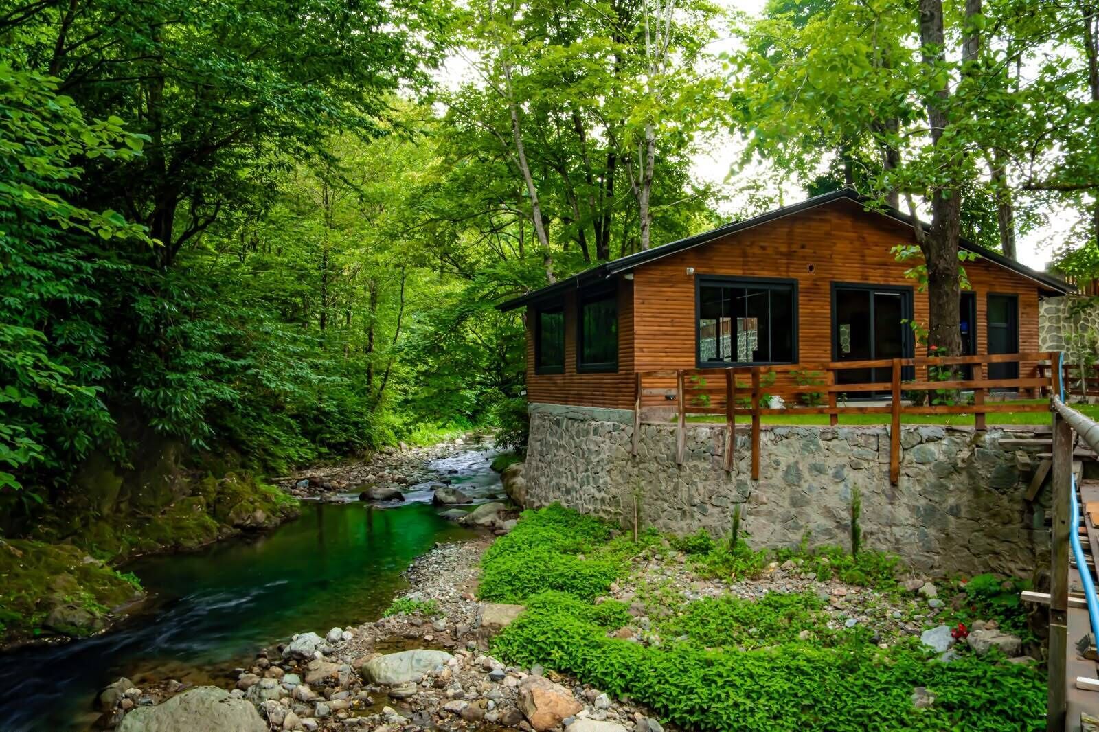 A wooden cabin with a stone foundation, surrounded by lush greenery and a flowing stream.
