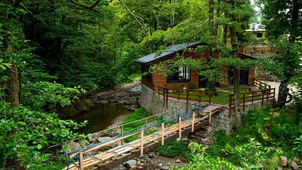 A rustic cabin with a wooden deck and a stone wall, surrounded by lush greenery and a flowing stream. - Dolana Wadi Taş Evleri (Ardeşen)
