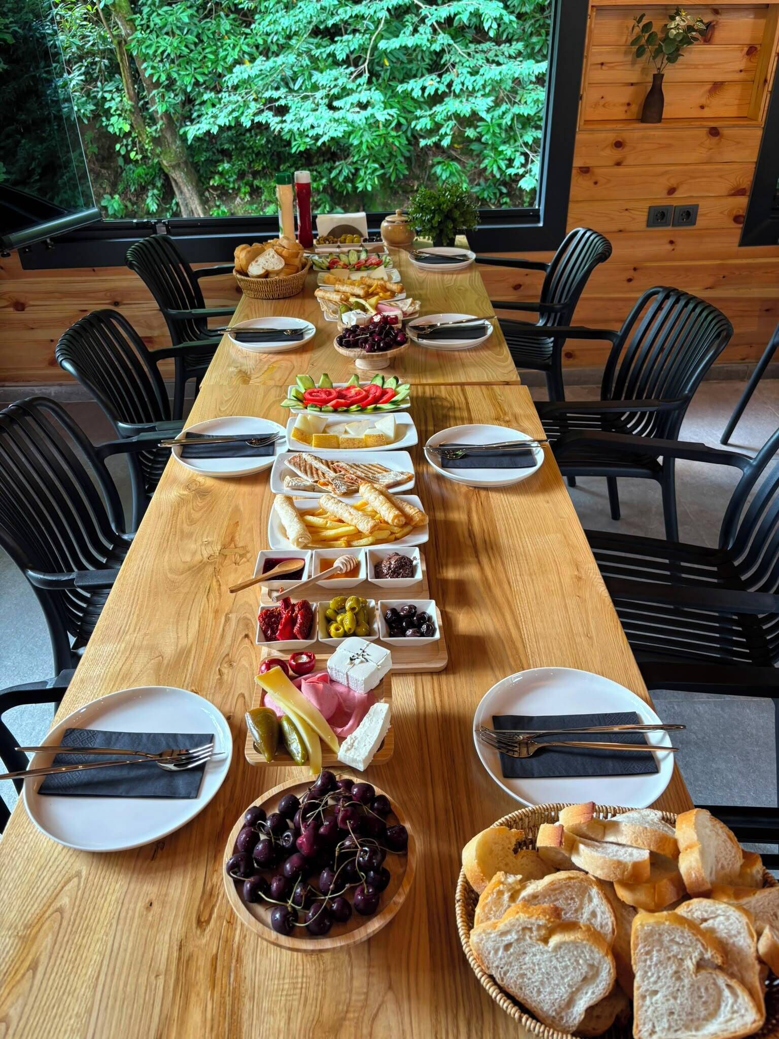 A long wooden dining table set for a meal with various dishes and bread, surrounded by black chairs, in a room with wooden walls and a view of greenery outside.