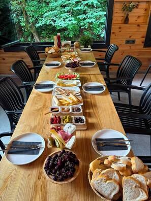 Uma longa mesa de jantar de madeira posta para uma refeição, com diversos pratos e pão, rodeada por cadeiras pretas, num espaço com paredes de madeira e vista para o verde lá fora.