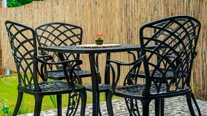 Juego de mesa y sillas de metal negro en un patio con baldosas, un camino tejido, un pequeño jarrón verde con flores y una valla de bambú al fondo.