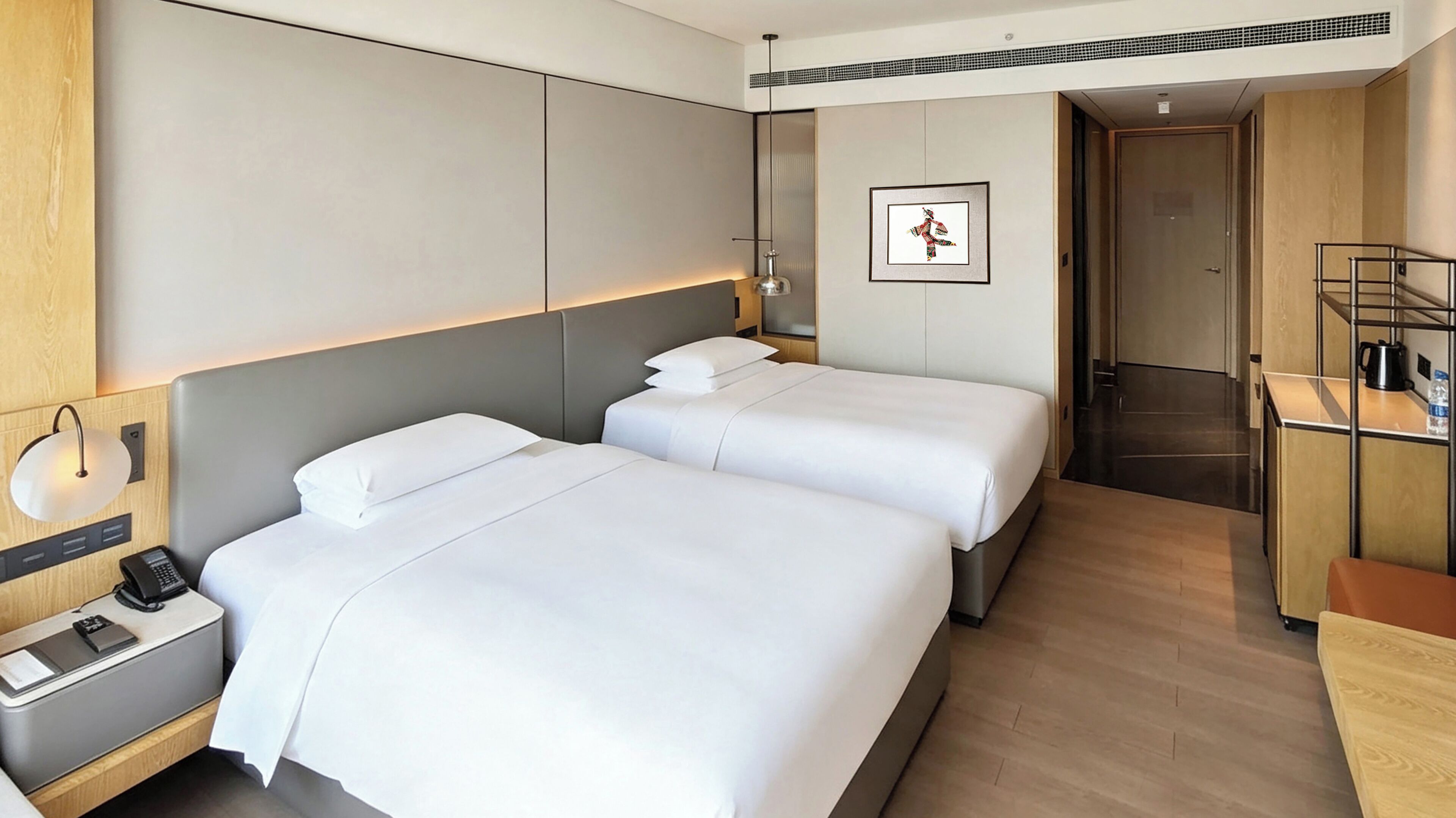 A hotel room with two beds, a bedside table with a phone, a wall-mounted light, and a framed picture on the wall.