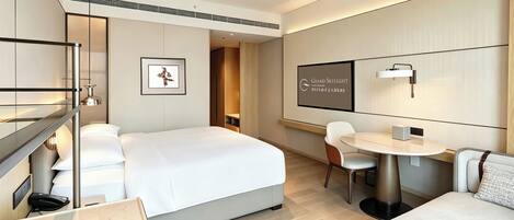 A modern hotel room with a large bed, a desk, a chair, and a small table. There is a framed picture on the wall and a sign with text.