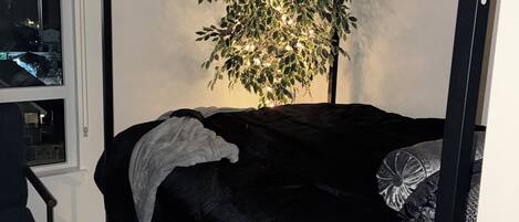 Un lit avec un cadre noir, un arbre décoré de guirlandes lumineuses, une fenêtre et une chaise.