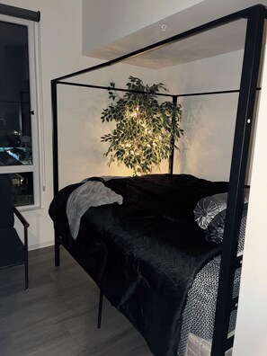 Ein Bett mit schwarzem Gestell, ein Baum mit Lichterketten, ein Fenster und ein Sessel.