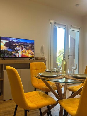 A dining area with a round glass table, yellow upholstered chairs, a television displaying a scenic view, and a door leading outside.