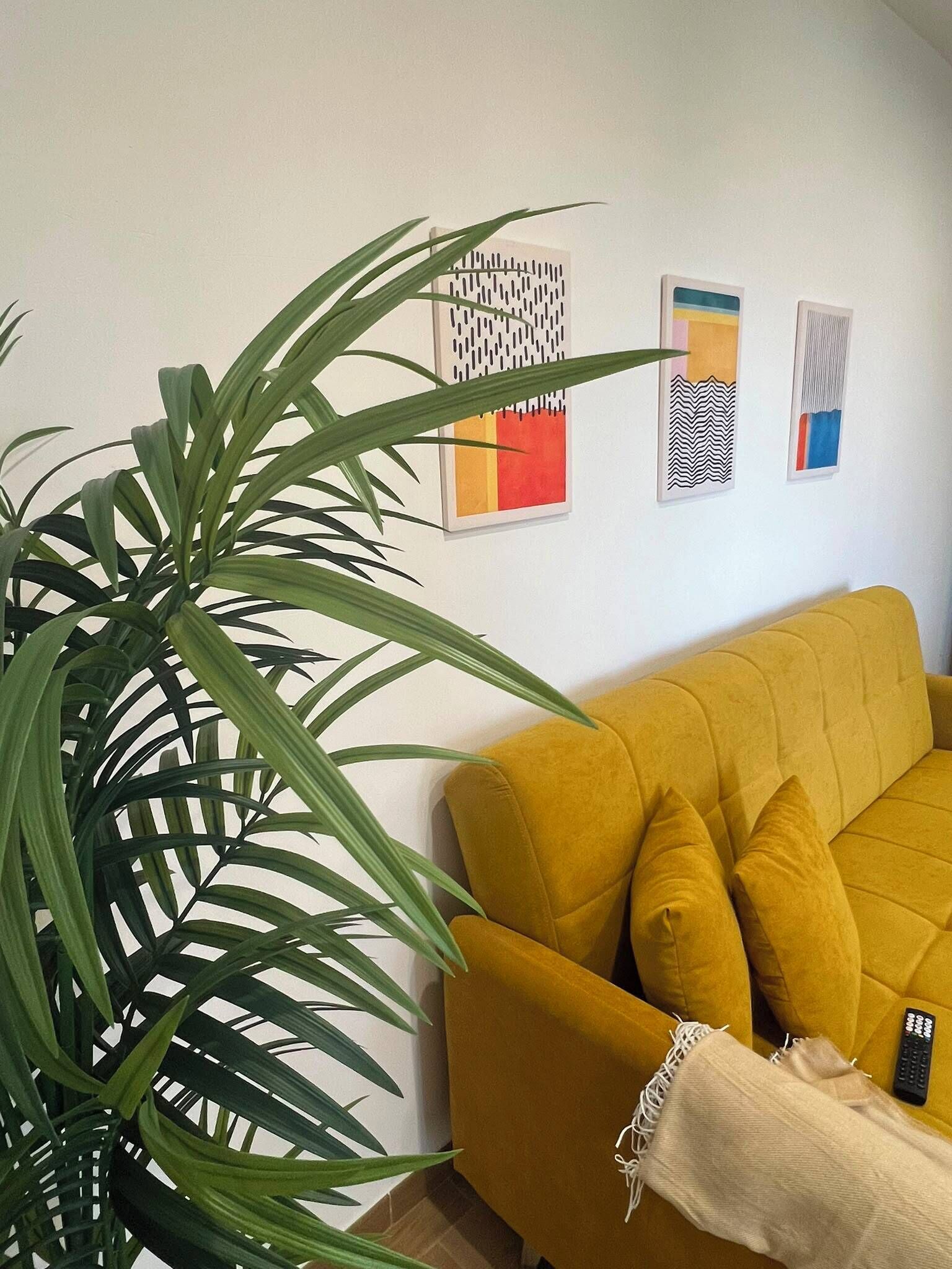 A living room with a yellow sofa, a large green plant, and three abstract wall art pieces.