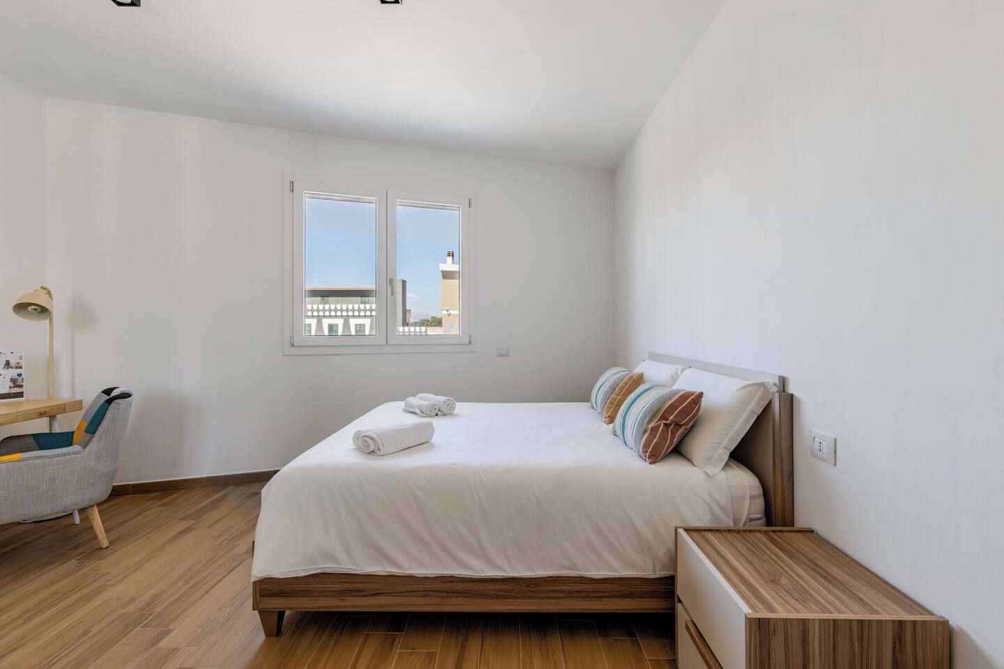 Een slaapkamer met een bed, een bureau, een stoel, een raam en uitzicht op gebouwen.