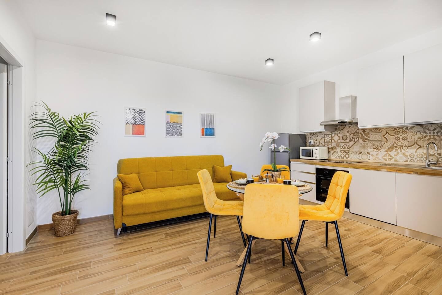 Ein stilvolles Wohnzimmer mit Sofa, Esstisch, Stühlen, einer Topfpflanze und einer Küchenzeile mit Mikrowelle.