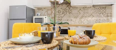 Une table de petit-déjeuner avec un plateau en verre, des chaises jaunes, une cafetière, une assiette de croissants et un verre de jus d’orange.