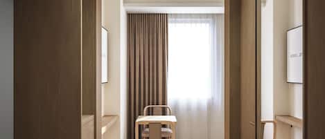 Habitación de hotel con piso de madera, un escritorio, una silla, un ventanal con cortinas y un televisor en la pared.