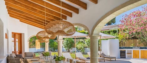 An outdoor dining area with a long table set for six, surrounded by wicker chairs, under a wooden pergola with hanging rattan lamps.