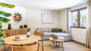 Ett modernt vardagsrum med en soffa, ett matbord med stolar, ett soffbord och ett litet sidobord. Det finns ett fönster med gardiner, ett konstverk som hänger på väggen och en dekorativ tallrik på väggen.