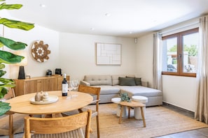 Ein modernes Wohnzimmer mit einer Couch, einem Esstisch mit Stühlen, einem Couchtisch und einem kleinen Beistelltisch. Ein Fenster mit Vorhängen, ein an der Wand befestigtes Kunstwerk und eine dekorative Platte sind zu sehen.