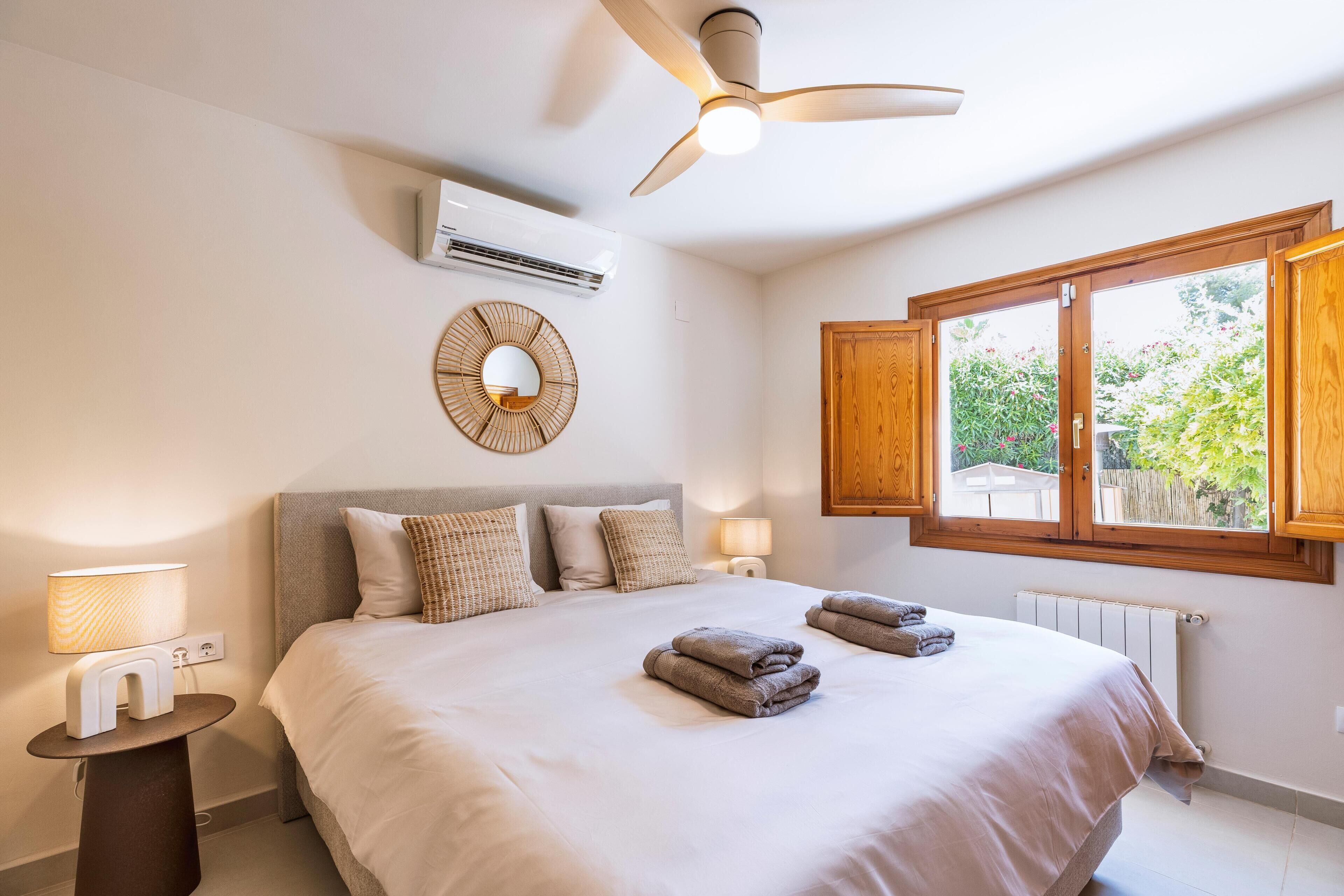 A bedroom with a bed, bedside lamps, a ceiling fan, an air conditioning unit, a window with wooden shutters, and a wall-mounted mirror.