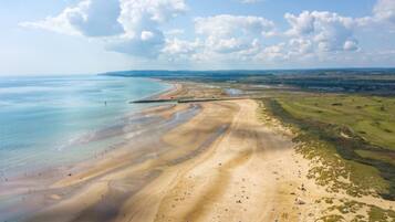 An aerial view of a coastal area with a sandy beach, a curved road, and distant green fields.