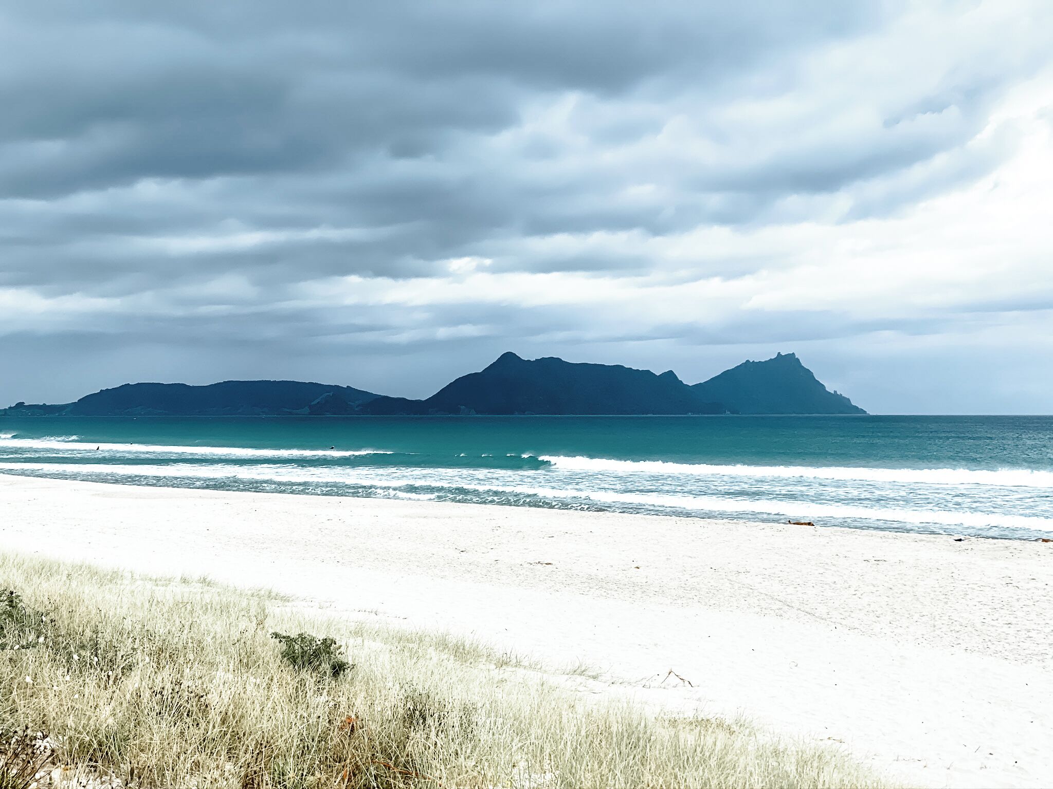 A beach with white sand and a view of mountains in the distance under a cloudy sky.