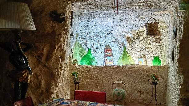 Un interior rústico con pared de piedra, una ventana con botellas de colores, una mesa con mantel estampado y una lámpara.