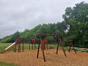 A playground with swings and a slide, surrounded by trees. - Cozy 3-Bed Chalet at Penlan Holiday Park | Pool (seasonal), WiFi & Dog Friendly (Cenarth)