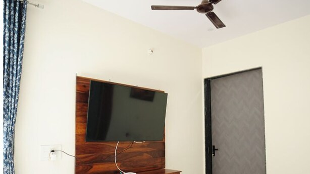A room with a flat-screen TV mounted on a wooden cabinet, a ceiling fan, and a door with a patterned door panel. - Luxury 3BHK Farmhouse Near Jaipur with Private Pool & Garden (jaipur)