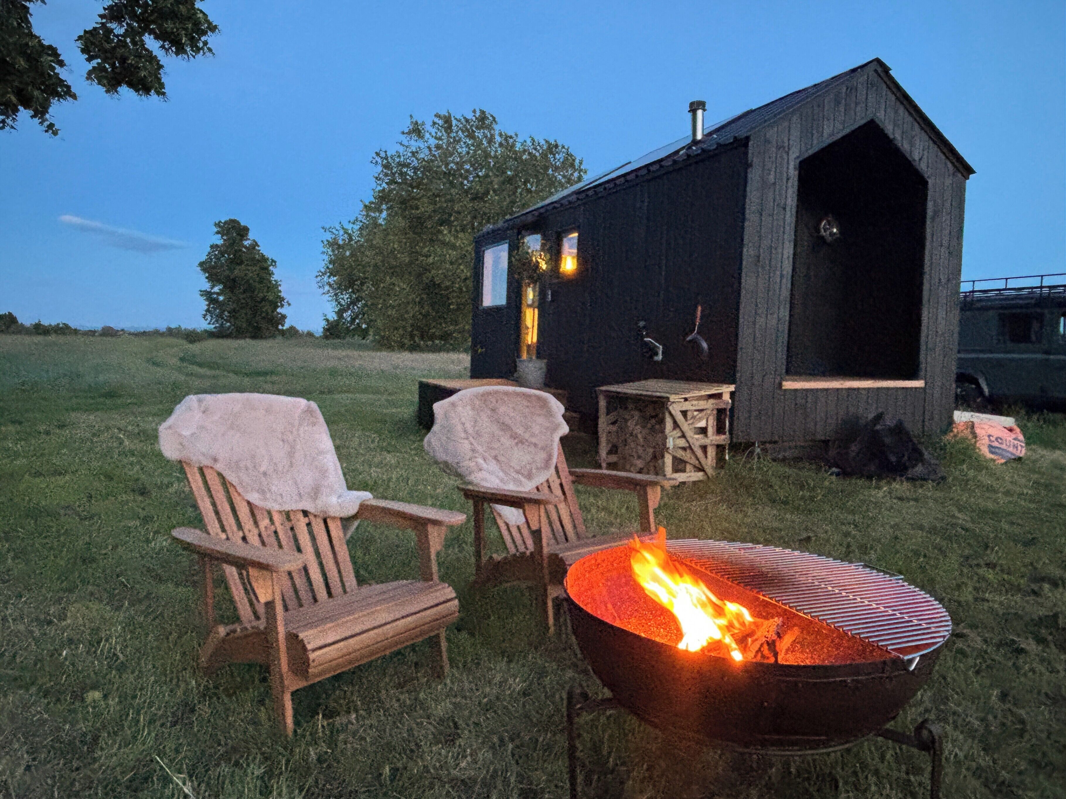 A rustic cabin with a fire pit, two covered chairs, and a small table.