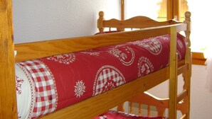Ein Stockbett mit gemustertem Bettbezug, ein Holzregal darüber mit verschiedenen Gegenständen und ein Fenster mit Vorhängen.