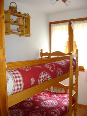 Uma cama beliche com colcha estampada, uma prateleira de madeira acima com vários objetos, e uma janela com cortinas.