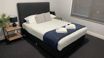 A neatly made bed with a dark headboard, white bedding, and a blue throw blanket. There are two bedside tables with lamps and a small plant on one.