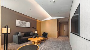 A modern hotel room with a sofa, a round table, a lamp, and a flat-screen TV mounted on the wall.