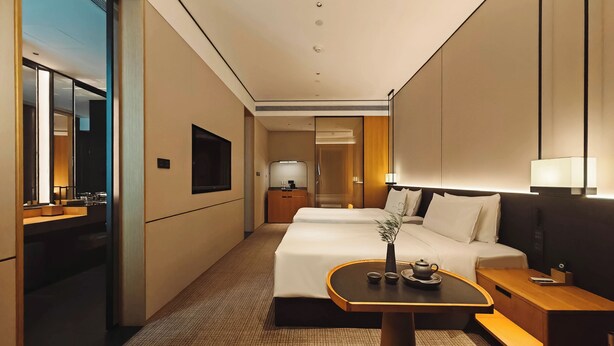 Habitación de hotel con cama, mesita de noche, lámpara y una pequeña mesa con un florero y tetera.