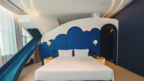 A modern hotel room with a large bed, a nightstand, a wall-mounted phone, and a control panel.