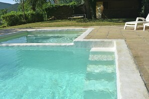 Ein rechteckiger Pool mit klarem Wasser, umgeben von einer Steindeck und einer Rasenfläche mit Bäumen und einer Bank.