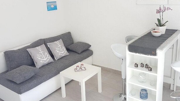 A compact living space with a grey sofa, a small white table, a white shelf unit, and a potted plant. - Apartments Katarina -one Bedroom Apartment With Terrace (Vis)