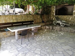 Eine Terrasse im Freien mit Steinboden, einem Tisch mit Stühlen, einer Bank und einem Grillbereich.