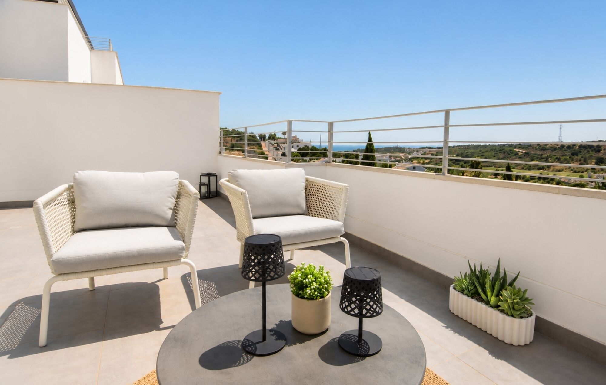 A rooftop terrace with two wicker chairs, a round table, two black lamps, and potted plants.