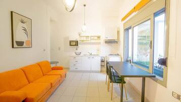 Un espacio habitable compacto con sofá, mesa de comedor, sillas, kitchenette y un ventanal grande.