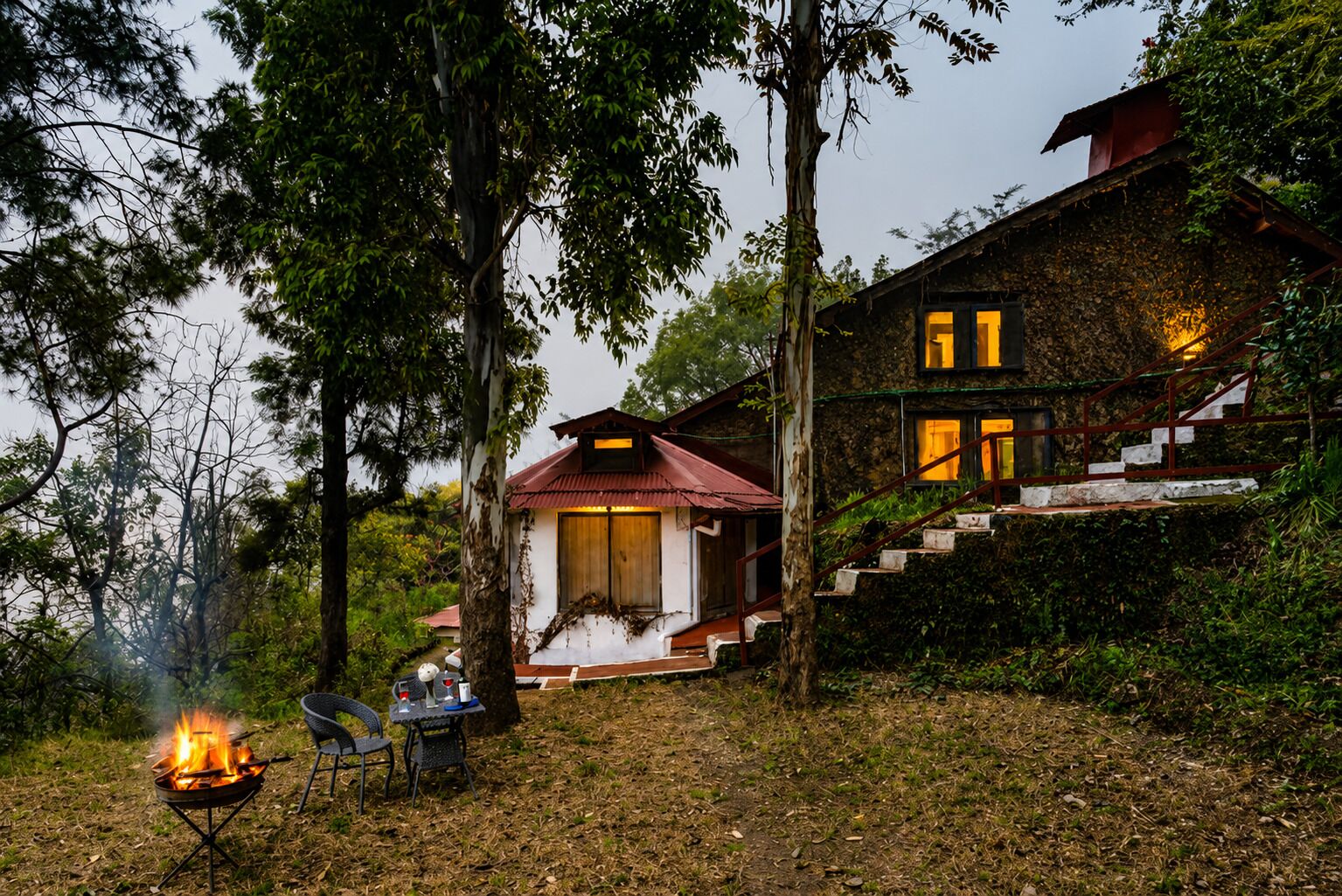 Una cabaña rústica con una fogata y asientos al aire libre, rodeada de árboles.