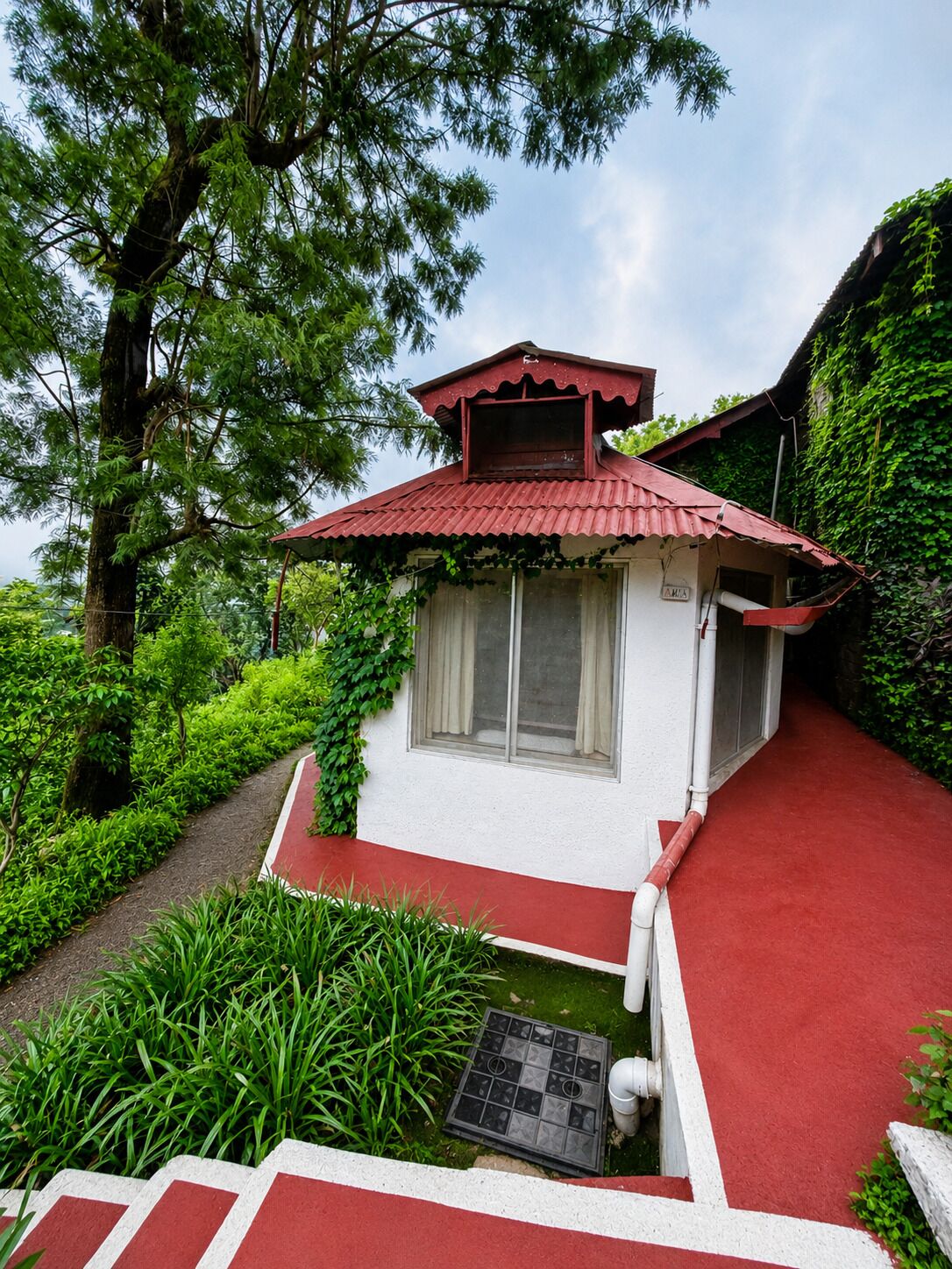 A small building with a red roof, surrounded by lush greenery and a red pathway leading to it.