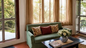 Un sofá verde con cojines estampados, una mesa de centro de madera con un libro y un jarrón de flores, y vistas a zonas verdes a través de grandes ventanales.