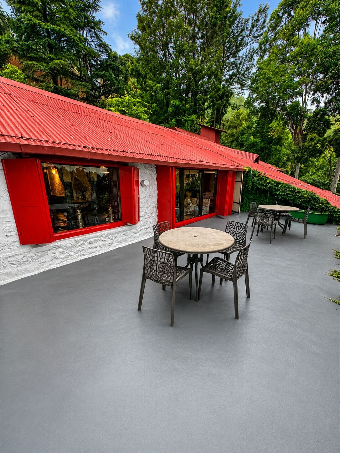 Un edificio con techo rojo y paredes blancas, con mesas redondas y sillas metálicas en su terraza, rodeado de vegetación.