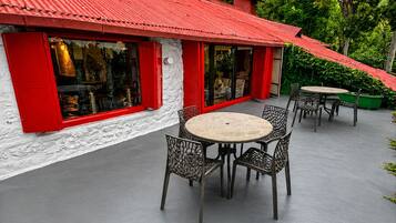 Un edificio con techo rojo y paredes blancas, con mesas redondas y sillas metálicas en su terraza, rodeado de vegetación.