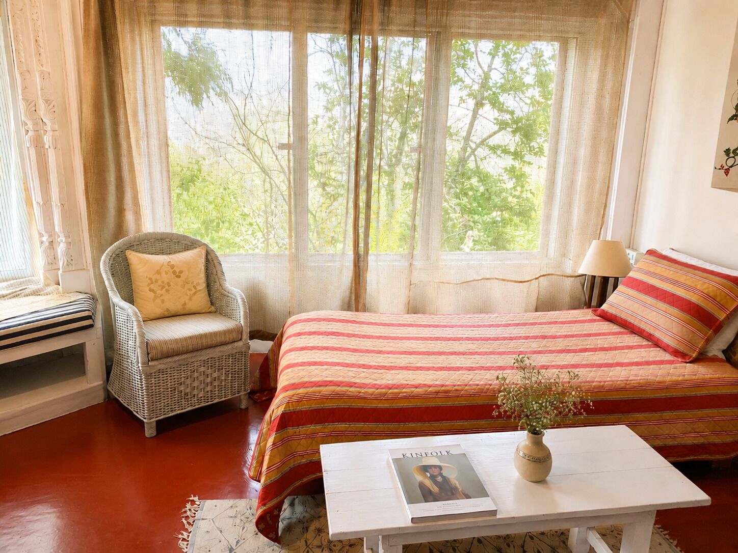 A bedroom with a large bed, a wicker chair, a bedside table with a book, and a window with curtains.
