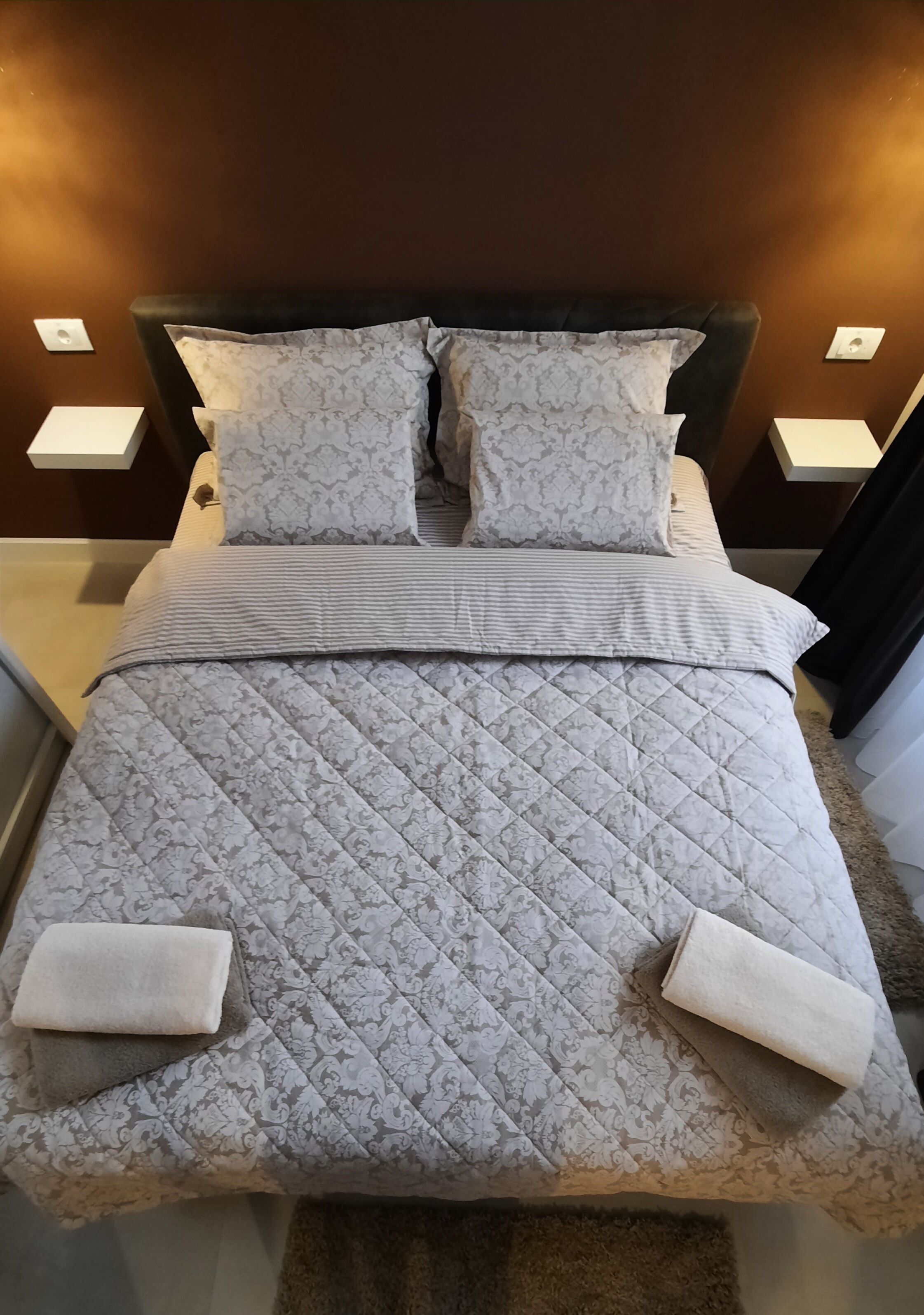 A bed with a quilted grey patterned bedspread, two matching pillows, and two folded towels on the footboard.