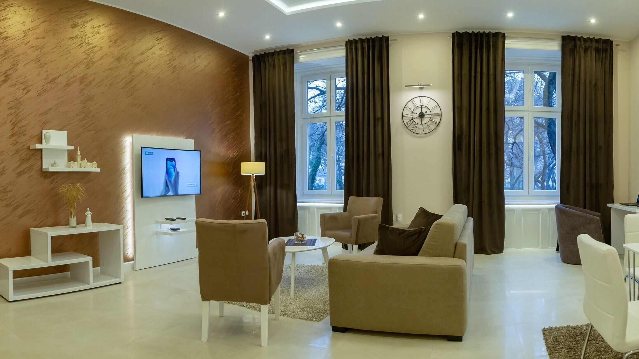 A modern living room with a flat-screen TV, a clock, and a view of trees through large windows.